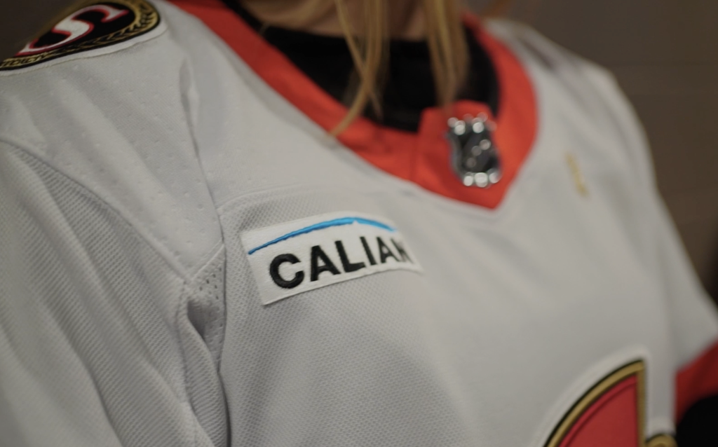Close-up of a white hockey jersey with a "CALIAN" patch on the shoulder and a red and black NHL logo near the collar.